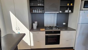 A compact kitchen with a gas stove, oven, and a built-in sink. There are cabinets with glassware and a refrigerator. - Blencathra Coastal Spa Getaway (Currie King Island)