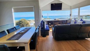 A spacious living area with a wooden floor, a dining table with chairs, a black sofa, and a TV mounted on the wall. - Blencathra Coastal Spa Getaway (Currie King Island)