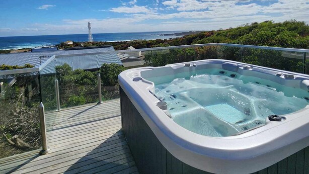 A hot tub on a wooden deck with a view of the ocean and a lighthouse in the distance. - Blencathra Coastal Spa Getaway (Currie King Island)