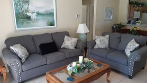 A living room with two grey sofas, a glass coffee table with decorative items, a painting of a landscape, and a kitchen area in the background.