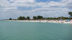 A beach with clear turquoise water, white sand, and a line of trees in the background.