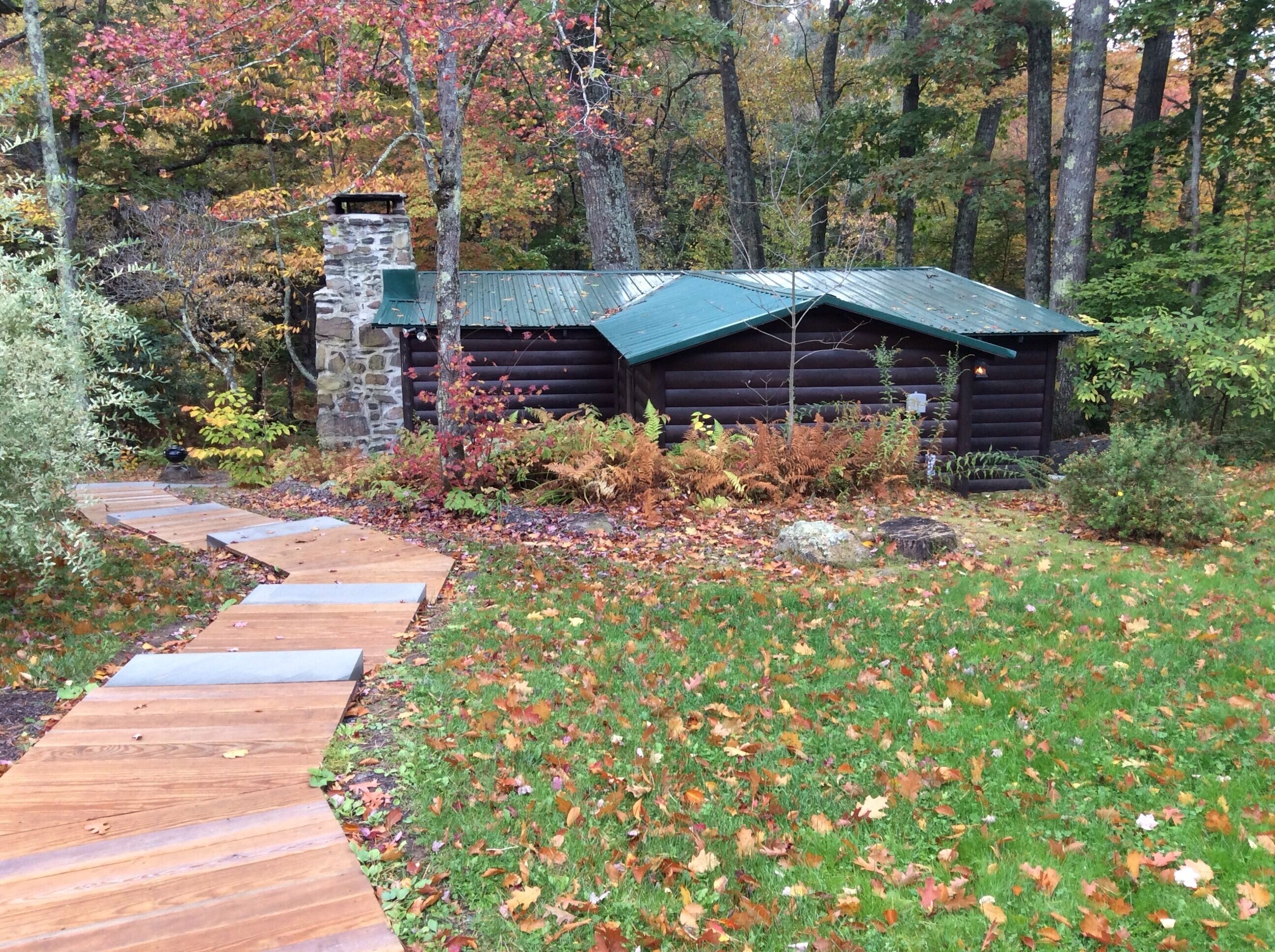 A rustic log cabin with a stone chimney, surrounded by a forest. A wooden pathway leads to the cabin.