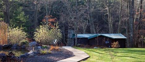 A rustic cabin with a wooden deck and a pond, surrounded by dense forest.