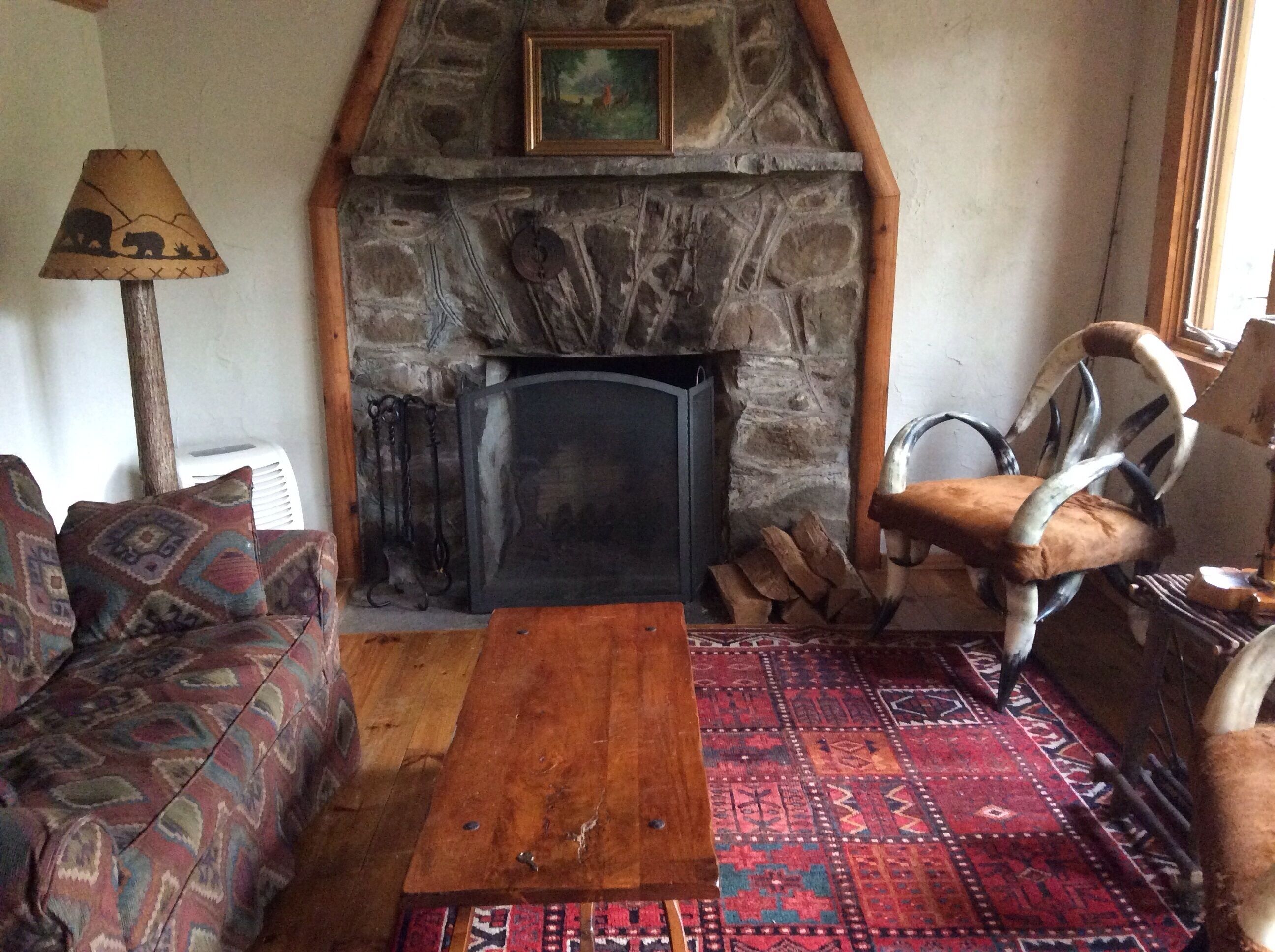 A cozy living room with a stone fireplace, a patterned sofa, a wooden coffee table, and a chair with antlers.