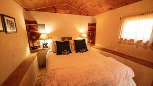 Een knus slaapkamer met een houten plafond, een groot bed met een witte dekbedovertrek, twee zwarte kussens en een nachtkastje met een lamp.
