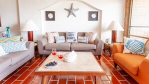 A living room with a grey sofa, an orange sofa, a coffee table with a decorative item and glasses, and a brick floor. - Playa Bonita Casa de Risa located on Los Muerto Beach  2BD Condo for rent in Los (Puerto Vallarta)