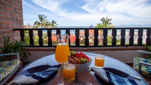 A table with a pitcher of orange juice, a bowl of fruit, and two glasses of orange juice on a balcony with a view of the sea and palm trees. - Playa Bonita Casa de Risa located on Los Muerto Beach  2BD Condo for rent in Los (Puerto Vallarta)