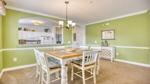 A dining room with a wooden table and white chairs, a kitchen in the background, and a green wall. - 325 River Crossing - Barefoot Resort Rentals (North Myrtle Beach)
