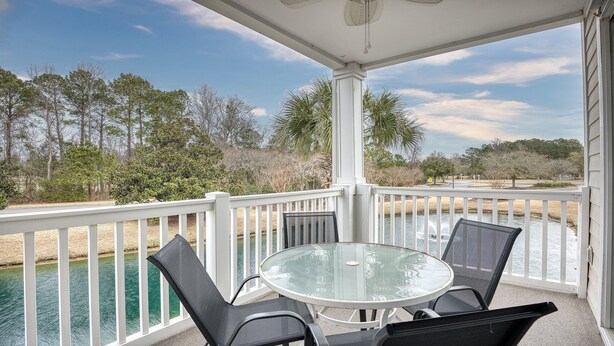 A covered outdoor area with a glass table and chairs, overlooking a body of water and surrounded by trees. - 325 River Crossing - Barefoot Resort Rentals (North Myrtle Beach)
