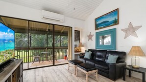 En rummelig opholdsstue med en stor skydedør, der fører til en balkon, en sort lædersofa, et sofabord og et fjernsyn på et træstel.