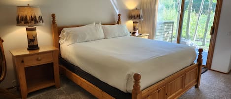 Une chambre à coucher avec un lit en bois, du linge de lit blanc, deux tables de chevet avec des lampes et une vue sur les arbres à travers une porte-fenêtre coulissante.