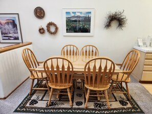 A dining area with a wooden table and chairs, a patterned rug, and wall decorations including a framed picture and two wreaths.