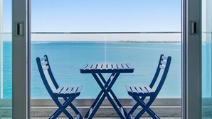 Un balcón con dos sillas plegables azules y una mesita, con vista a un mar tranquilo y una costa lejana.