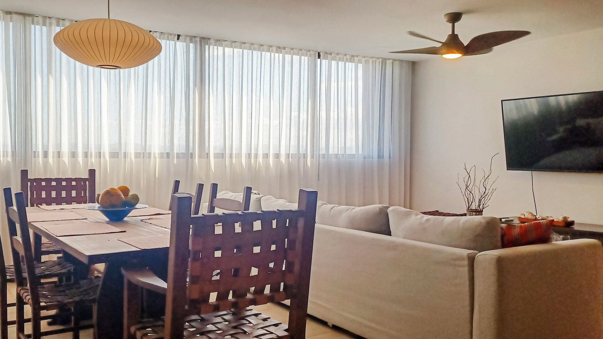 A living room with a sofa, dining table, chairs, a ceiling fan, and a TV mounted on the wall.