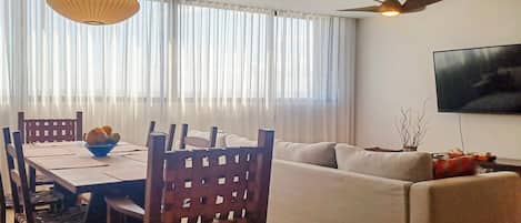 A living room with a sofa, dining table, chairs, a ceiling fan, and a TV mounted on the wall.