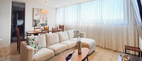 A living room with a sofa, a coffee table, a dining table with chairs, and a large window with curtains.