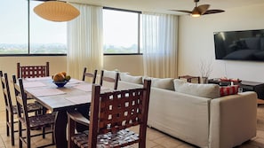 A modern living room with a dining area, large windows, a ceiling fan, and a flat-screen TV.