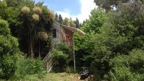 Una casa de madera rústica rodeada de abundante vegetación, incluyendo palmeras y diversos arbustos. Una escalera conduce a una puerta roja, y hay una pila de leña cerca.