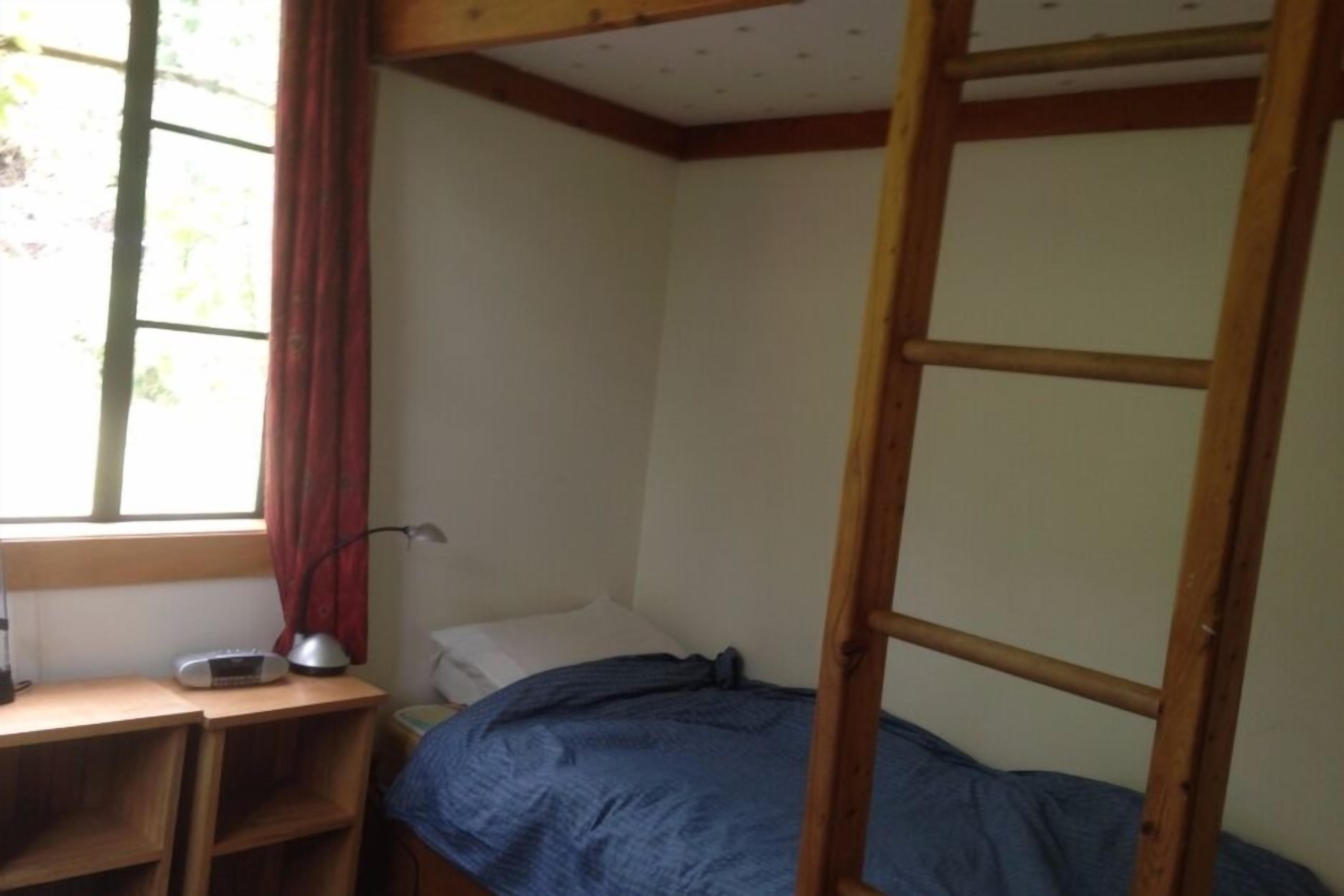 A small, single-bed room with a wooden bunk bed, a desk with a lamp, and a window with curtains.