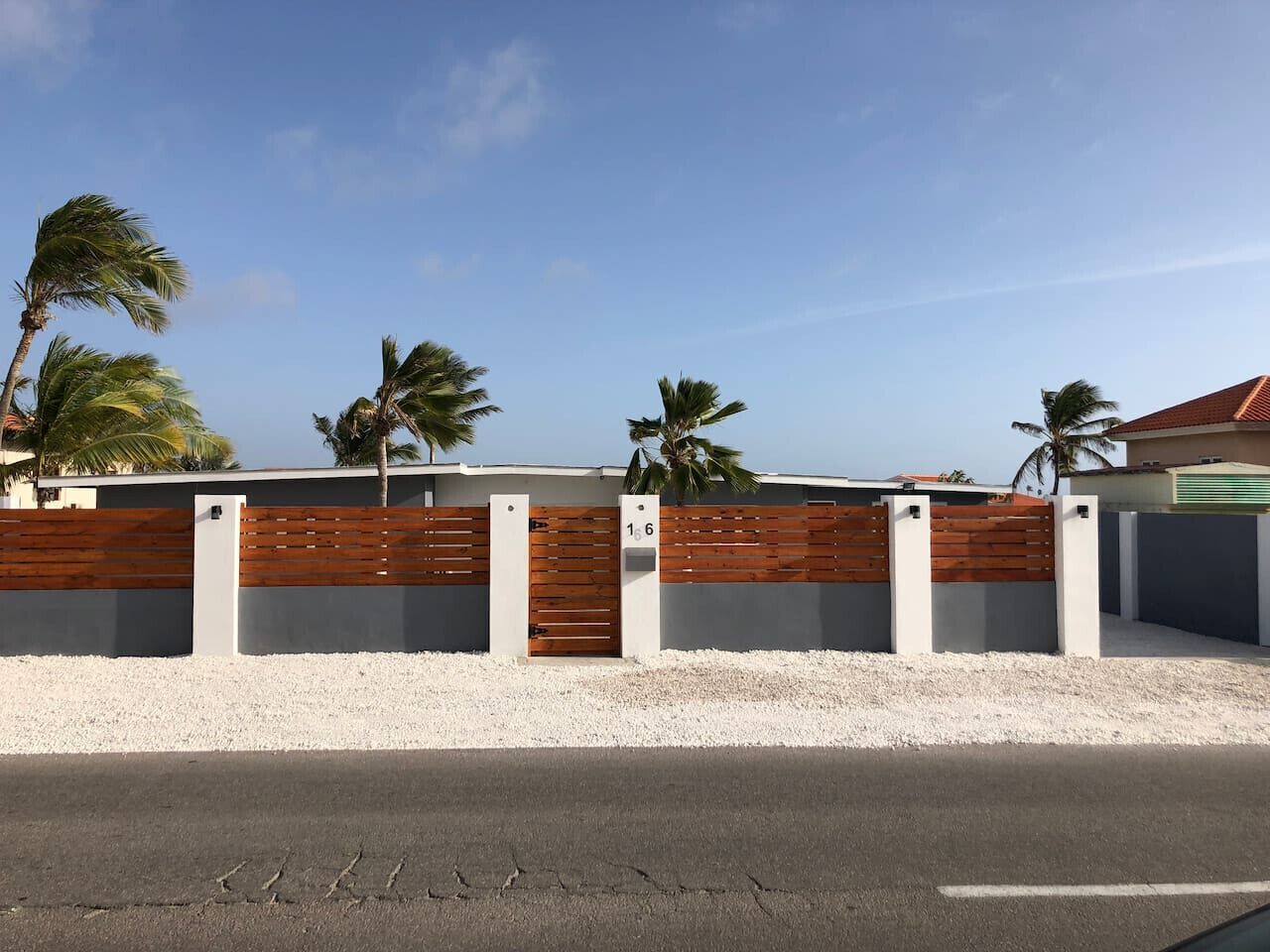 Een modern gebouw met een houten omheining en poort, palmbomen op de achtergrond, en een heldere lucht.