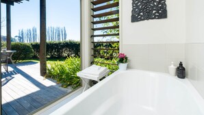Ein Badezimmer mit einer Badewanne, einem Fenster mit Holzjalousien, einer Wanddekoration und einem kleinen Tisch mit einem Handtuch.