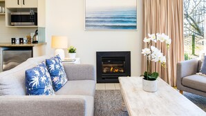 Ein Wohnzimmer mit grauem Sofa, einem Couchtisch, einem Kamin und einer Küchenzeile, mit Blick auf das Meer durch ein Fenster.