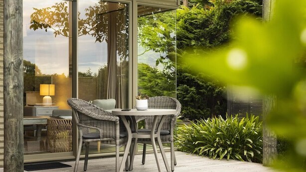 Eine Terrasse mit Korbmöbeln und einem Tisch, umgeben von üppigem Grün und einer Glastür, die zu einem Raum mit einer Lampe führt.