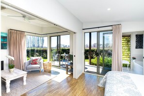 A modern bedroom with a large sliding glass door leading to a balcony, a bed with a patterned bedspread, a small table with a plant, and a chair with a red pillow.