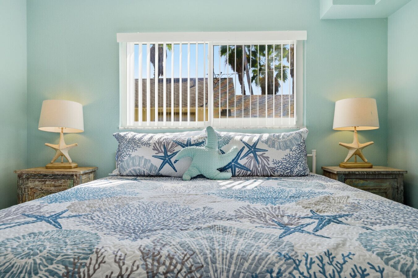 A bedroom with a bed featuring a blue and white coral pattern, two starfish lamps, and a window with a view of palm trees and a brick building.