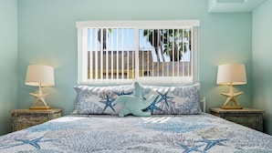 A bedroom with a bed featuring a blue and white coral pattern, two starfish lamps, and a window with a view of palm trees and a brick building.