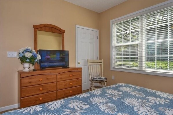 Un dormitorio con cama, cómoda de madera, televisor, silla, ventana con persianas y puerta.