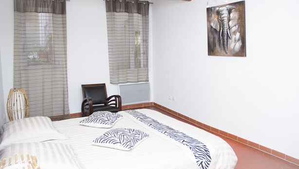 Ein Schlafzimmer mit einem Bett, zwei Vorhängen an den Fenstern, einem Sessel und einem Wandbild.