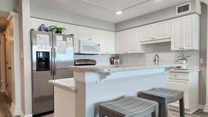 A modern kitchen with white cabinetry, a stainless steel refrigerator, a microwave, and a built-in oven. There are two bar stools and a small island with a sink. - Crescent C4B - Nicely Furnished - Oceanfront - Private Balcony (Pawleys Island)