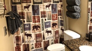 Un baño con cortina de ducha a cuadros con motivos de fauna y cabañas, un perchero con una toalla enrollada y una encimera de granito con un lavabo.
