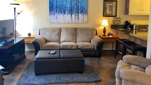 Una sala de estar con sofá, reposapiés y un cuadro de un bosque. Se observa un televisor, una lámpara y una mesita con una lámpara. En el fondo, se distingue una zona de cocina.