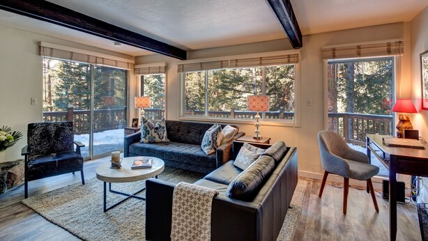 A cozy living room with a sofa, armchairs, and a coffee table. Large windows offer a view of the outdoors.