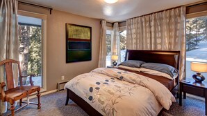 A bedroom with a large bed, a chair, a bedside table with a lamp, and a window with curtains.