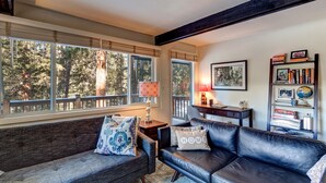A cozy living room with a dark leather sofa, a grey couch, and a wooden coffee table. There are large windows with a view of trees outside.