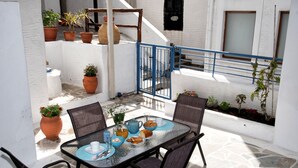 Un área de comedor al aire libre iluminada por el sol, con una mesa para dos, rodeada de plantas en macetas y un portón azul.