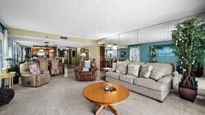 Un salon spacieux avec un canapé beige, un fauteuil en cuir marron et une table basse en bois. On aperçoit, au fond, une grande plante en pot et un coin repas.