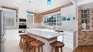 Cozinha moderna com ilha grande, armários brancos, parede de tijolos e janela com vista para o verde.