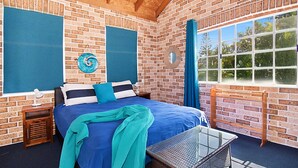 Quarto com parede de tijolos, cama com roupa de cama azul, mesa de cabeceira, abajur, janela com vista para árvores e cortina azul.