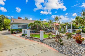 A residential house with a paved driveway, a fenced garden area with palm trees, and a red car parked in the driveway.