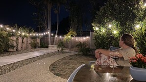 Une personne assise à une table, un verre à la main, à l’extérieur, de nuit, avec des guirlandes lumineuses et des plantes.