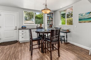 A dining area with a round table, four chairs, a bookshelf, and a window with a view of greenery. - Relaxing, Romantic, and Fun! Private, Quiet, and Less than a mile to the Ocean! (Oceanside)