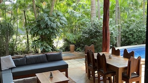 Een overdekt terras met een bank, eettafel en stoelen, omgeven door weelderige begroeiing en een zwembad.