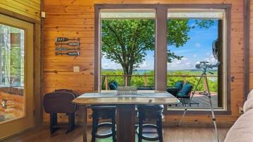 A wooden cabin interior with a dining table and chairs, a telescope on the balcony, and a view of a tree and landscape.