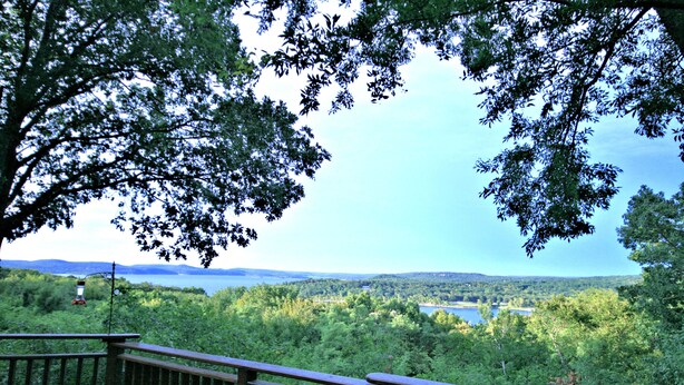 Une terrasse en bois, dotée d’une balustrade, surplombe un lac entouré d’une forêt dense et de collines.
