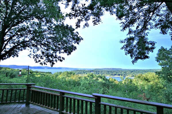 Une terrasse en bois, dotée d’une balustrade, surplombe un lac entouré d’une forêt dense et de collines.