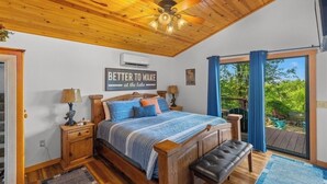 A bedroom with a wooden ceiling, a bed with a headboard, a bench, a nightstand, a lamp, and a window with curtains.